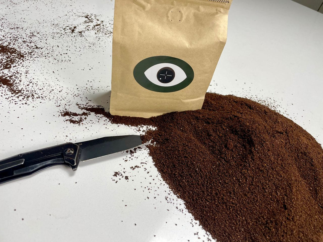 Bag with an eye design labeled 'DARK ARTS' next to a pile of ground coffee and a black knife on a white surface.