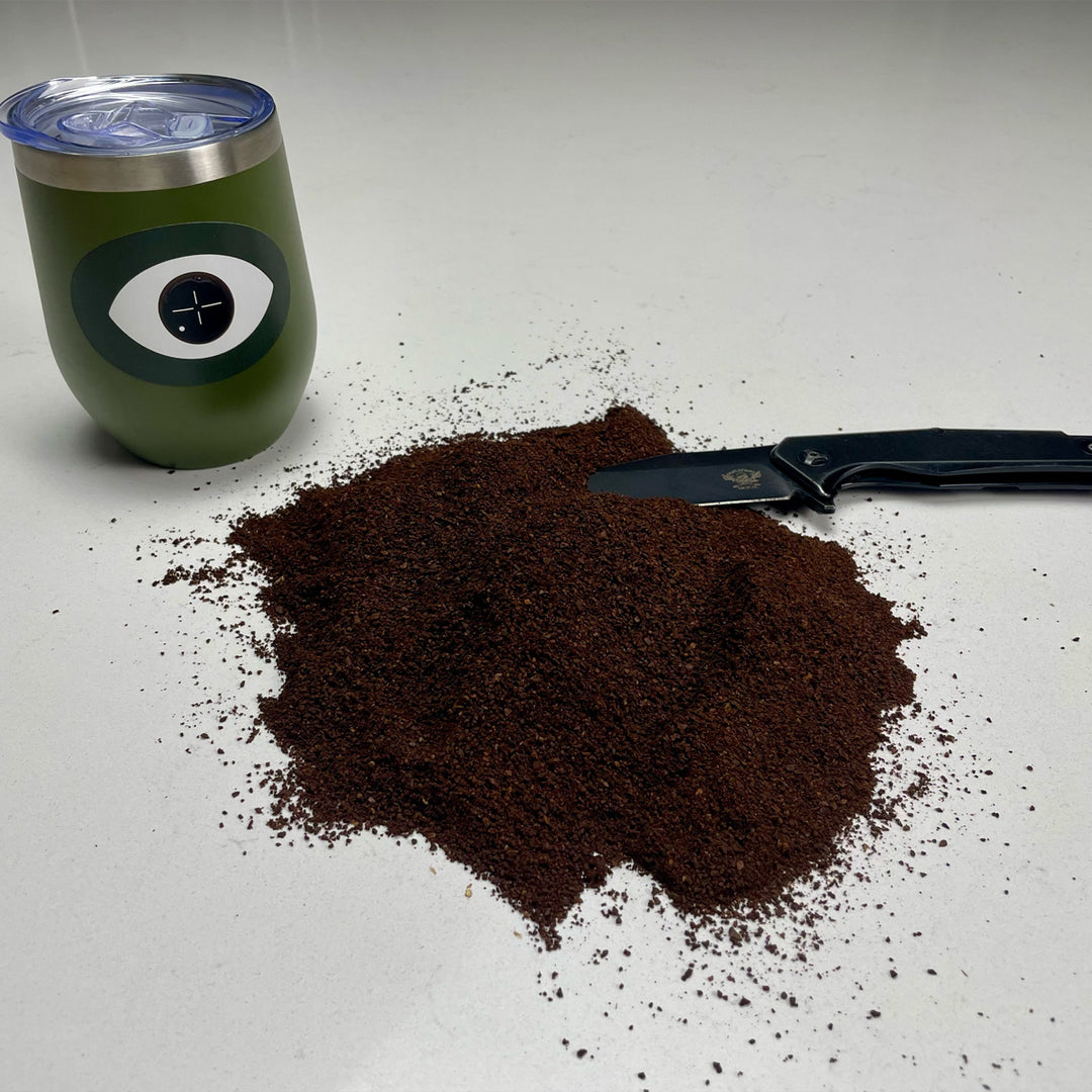 Coffee grounds spilled on the surface next to a coffee tumbler with a Dark Arts logo and a knife.