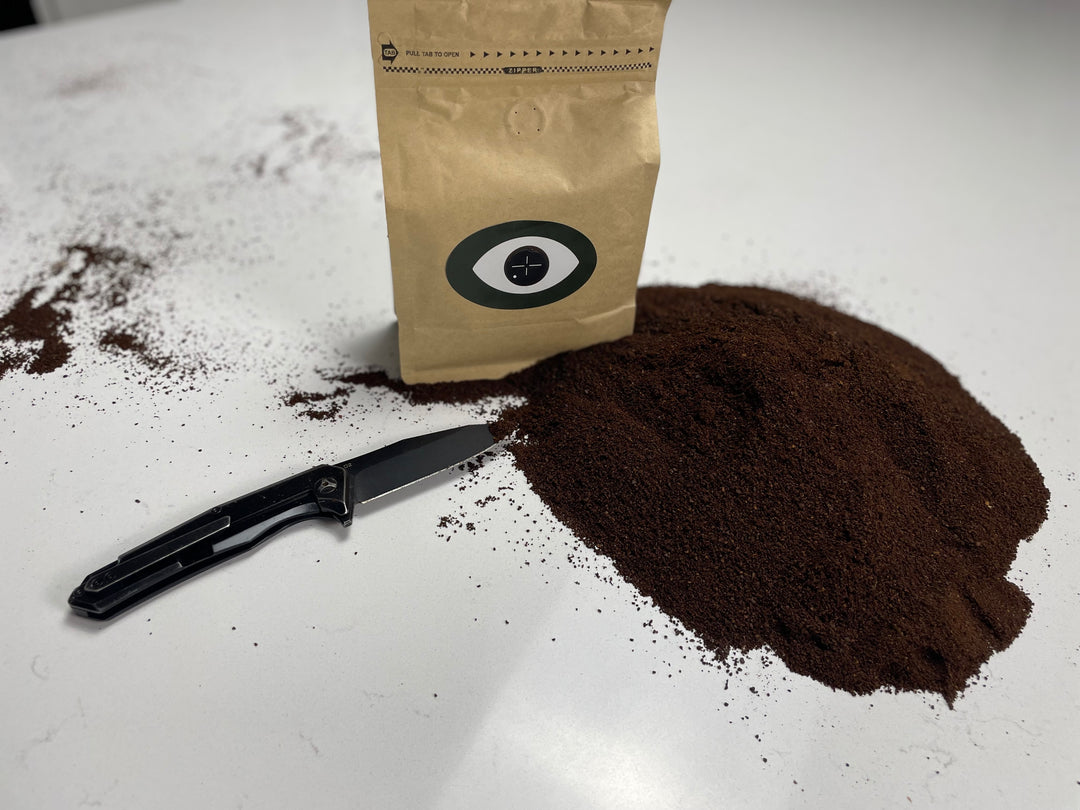 Ground coffee spilled on a white surface next to a brown Dark Arts coffee bag and a black knife.