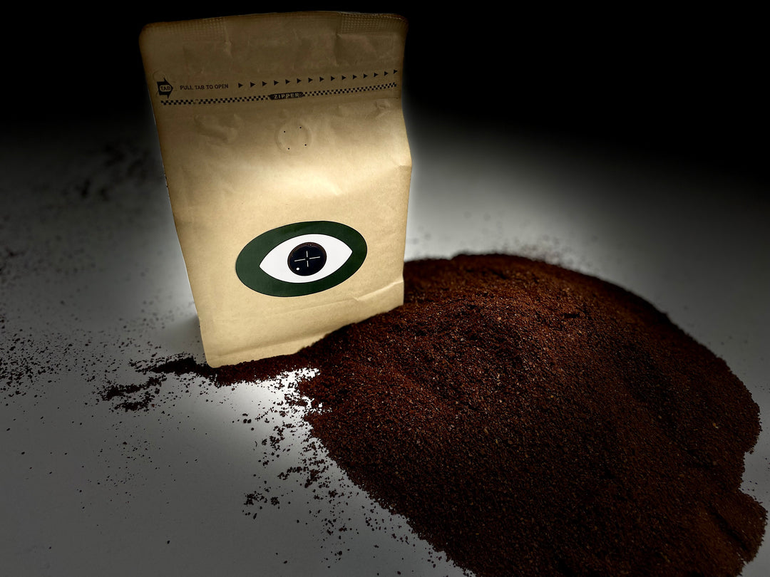 Moody spotlight image of a brown coffee bag with a green eye logo, placed next to a large mound of ground coffee on a dark background with scattered coffee grounds.