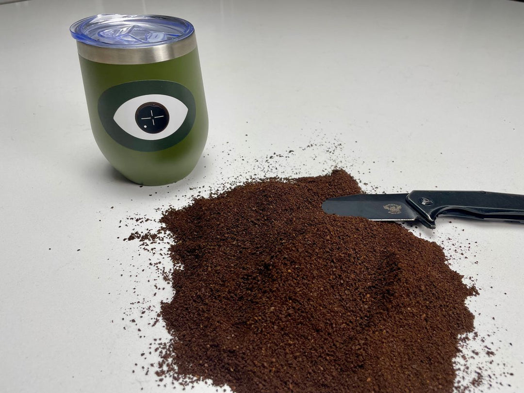 Green tumbler with an eye design next to a pile of ground coffee and a black knife on a white surface.